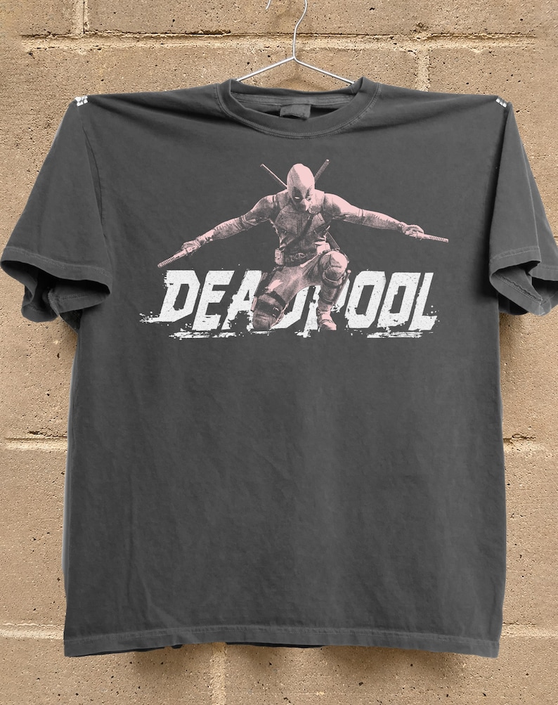 Puede incluir: Camiseta gris oscuro con un gr&aacute;fico de Deadpool en posici&oacute;n de combate, sosteniendo dos espadas. La palabra "DEADPOOL" est&aacute; impresa en blanco, con letras desgastadas en la parte inferior de la imagen.