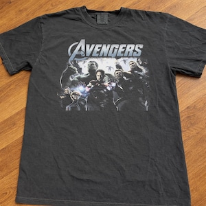May include: Dark gray t-shirt featuring the Avengers team. The shirt displays the Avengers logo in white and blue, with a graphic of the characters. The shirt is made of a soft material.