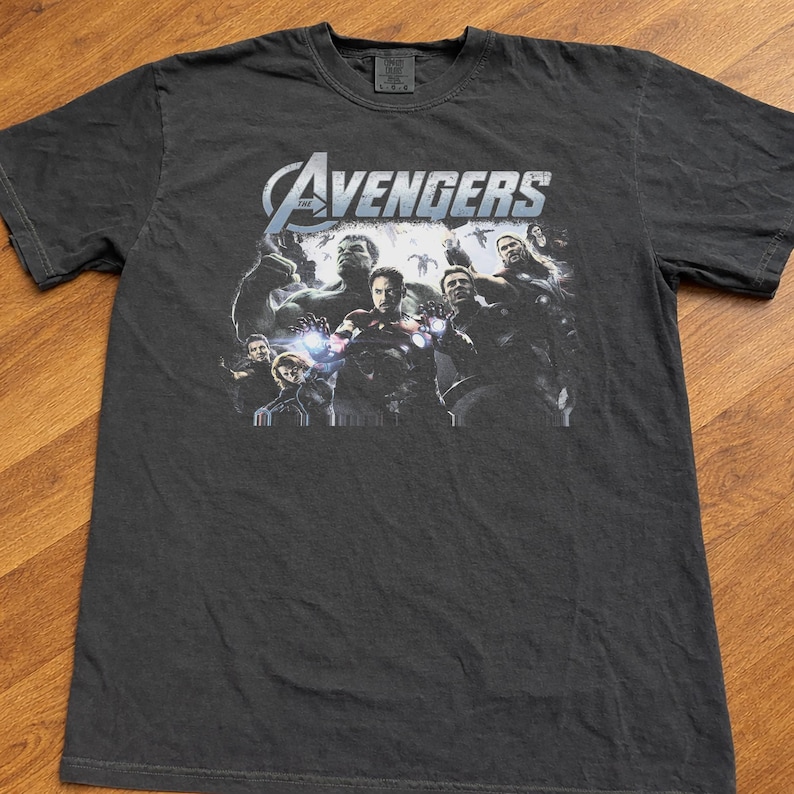 May include: Dark gray t-shirt featuring the Avengers team. The shirt displays the Avengers logo in white and blue, with a graphic of the characters. The shirt is made of a soft material.