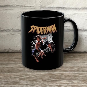 Puede incluir: Una taza de cer&aacute;mica negra con un gr&aacute;fico del personaje Spider-Man en una pose din&aacute;mica. Sobre el personaje, la palabra SPIDER-MAN est&aacute; impresa en una fuente amarilla estilizada y dentada. El dise&ntilde;o incluye un fondo de ciudad en blanco y negro.
