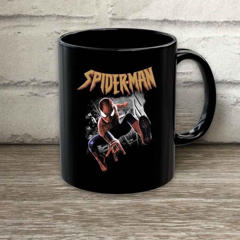 Puede incluir: Una taza de cer&aacute;mica negra con un gr&aacute;fico del personaje Spider-Man en una pose din&aacute;mica. Sobre el personaje, la palabra SPIDER-MAN est&aacute; impresa en una fuente amarilla estilizada y dentada. El dise&ntilde;o incluye un fondo de ciudad en blanco y negro.