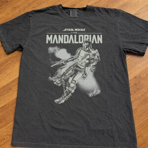 Op de afbeelding: Donkergrijs t-shirt met een afbeelding van The Mandalorian uit Star Wars. Het ontwerp bevat de tekst "STAR WARS THE MANDALORIAN" boven een afbeelding van het personage. Het shirt is gemaakt van een zacht materiaal.