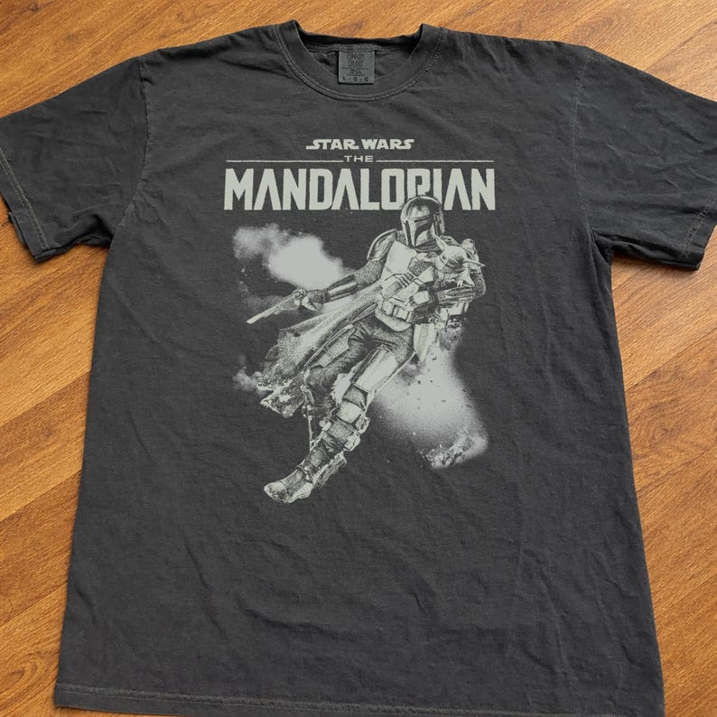 Op de afbeelding: Donkergrijs t-shirt met een afbeelding van The Mandalorian uit Star Wars. Het ontwerp bevat de tekst "STAR WARS THE MANDALORIAN" boven een afbeelding van het personage. Het shirt is gemaakt van een zacht materiaal.
