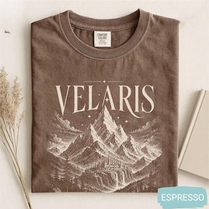 Pode incluir: Uma t-shirt castanha de gola redonda com um gráfico em cor creme que mostra uma cordilheira e uma cidade na base. O texto VELARIS está impresso numa fonte serifada grande sobre a ilustração da montanha, decorada com pequenas estrelas.