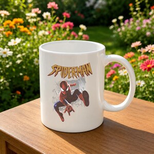 Puede incluir: Una taza de cer&aacute;mica blanca con un gr&aacute;fico del superh&eacute;roe Spider-Man en una pose din&aacute;mica agachada. Sobre el personaje, la palabra SPIDER-MAN est&aacute; impresa en una fuente amarilla estilizada y dentada con un contorno oscuro sobre un fondo gris.