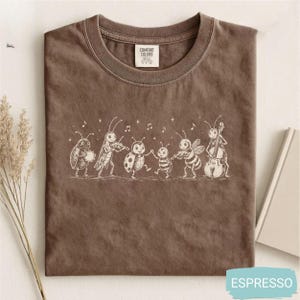 Puede incluir: Una camiseta marrón de cuello redondo con una ilustración de líneas blancas de un grupo de insectos tocando instrumentos musicales, incluyendo pandereta, violín y violonchelo, con notas musicales flotando y una etiqueta de Comfort Colors.