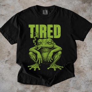 Pode incluir: Uma t-shirt preta de manga curta com um gráfico de um sapo verde com um cigarro na boca e a palavra TIRED impressa em letras verdes grandes acima do sapo. O sapo tem uma aparência texturizada e detalhada com patas palmadas visíveis.