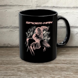Puede incluir: Una taza de cer&aacute;mica negra con un gr&aacute;fico en rosa claro del personaje Spider-Man en una pose agachada. El texto SPIDER-MAN est&aacute; impreso en rosa claro sobre el personaje. El dise&ntilde;o incluye patrones abstractos de telara&ntilde;a en el fondo.