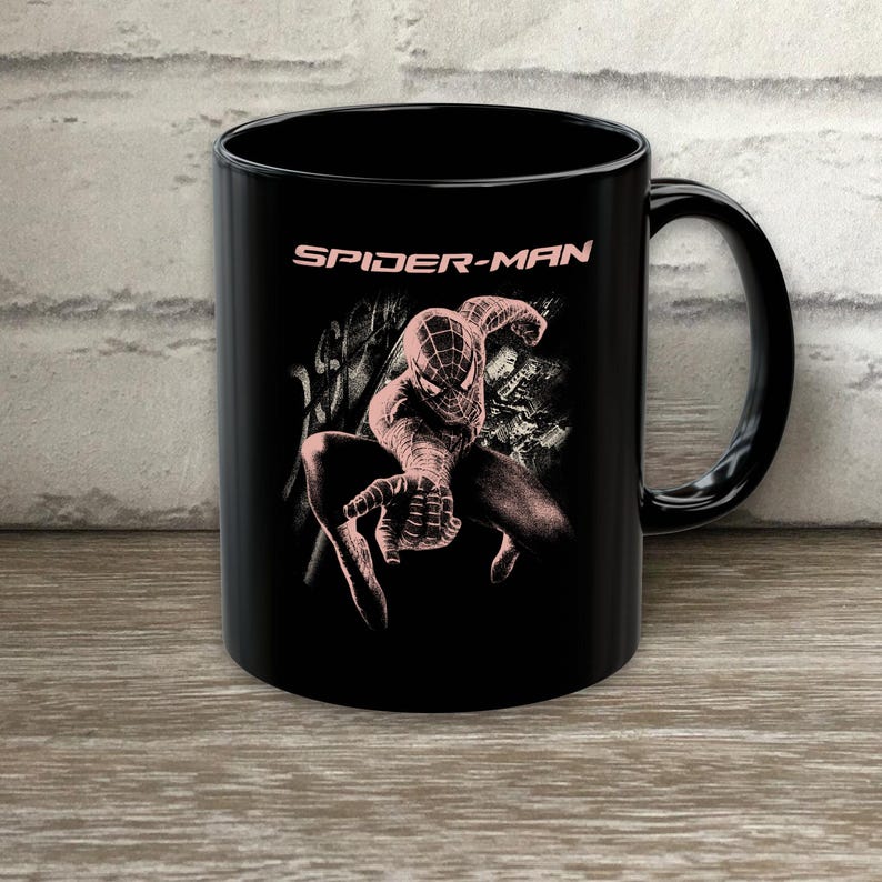 Puede incluir: Una taza de cer&aacute;mica negra con un gr&aacute;fico en rosa claro del personaje Spider-Man en una pose agachada. El texto SPIDER-MAN est&aacute; impreso en rosa claro sobre el personaje. El dise&ntilde;o incluye patrones abstractos de telara&ntilde;a en el fondo.