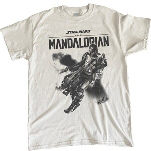 Op de afbeelding: Wit t-shirt met de zwarte tekst "STAR WARS THE MANDALORIAN". Daaronder een zwart-wit afbeelding van het Mandalorian-personage, met een blaster en in actie.