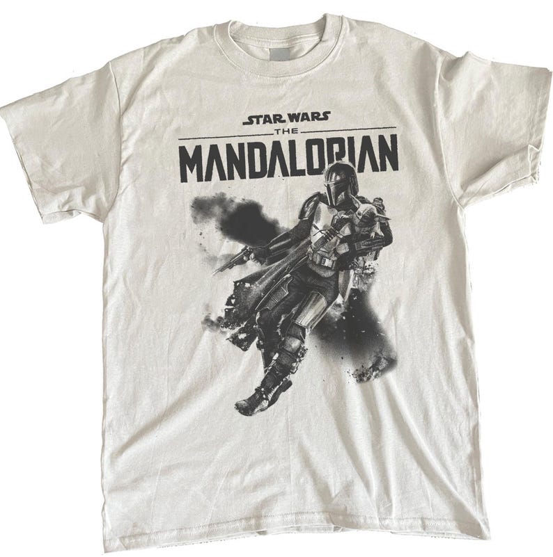 Op de afbeelding: Wit t-shirt met de zwarte tekst "STAR WARS THE MANDALORIAN". Daaronder een zwart-wit afbeelding van het Mandalorian-personage, met een blaster en in actie.