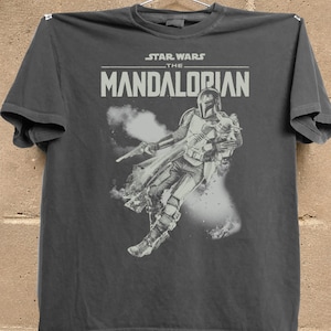 Op de afbeelding: Donkergrijs t-shirt met een afbeelding van The Mandalorian uit Star Wars. Het ontwerp bevat de titel van de show en een gedetailleerde illustratie van het personage in actie, met een blaster en cape.