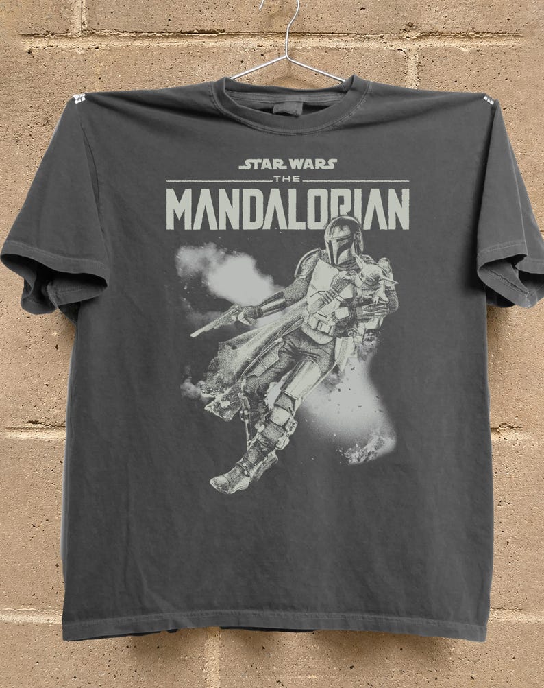 Op de afbeelding: Donkergrijs t-shirt met een afbeelding van The Mandalorian uit Star Wars. Het ontwerp bevat de titel van de show en een gedetailleerde illustratie van het personage in actie, met een blaster en cape.