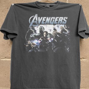 May include: Dark gray t-shirt featuring the text "The Avengers" in white and blue, with a graphic of several Avengers characters. The shirt is hanging on a hanger against a brick wall.