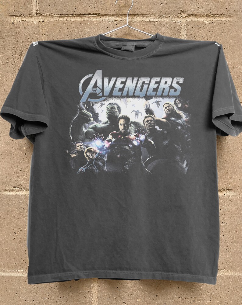 May include: Dark gray t-shirt featuring the text "The Avengers" in white and blue, with a graphic of several Avengers characters. The shirt is hanging on a hanger against a brick wall.