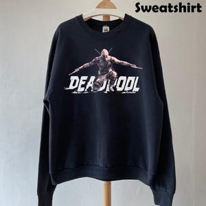 Puede incluir: Sudadera negra con un gr&aacute;fico de Deadpool y la palabra "DEADPOOL" en letras blancas desgastadas. La parte superior de manga larga est&aacute; colgada en una percha de madera.