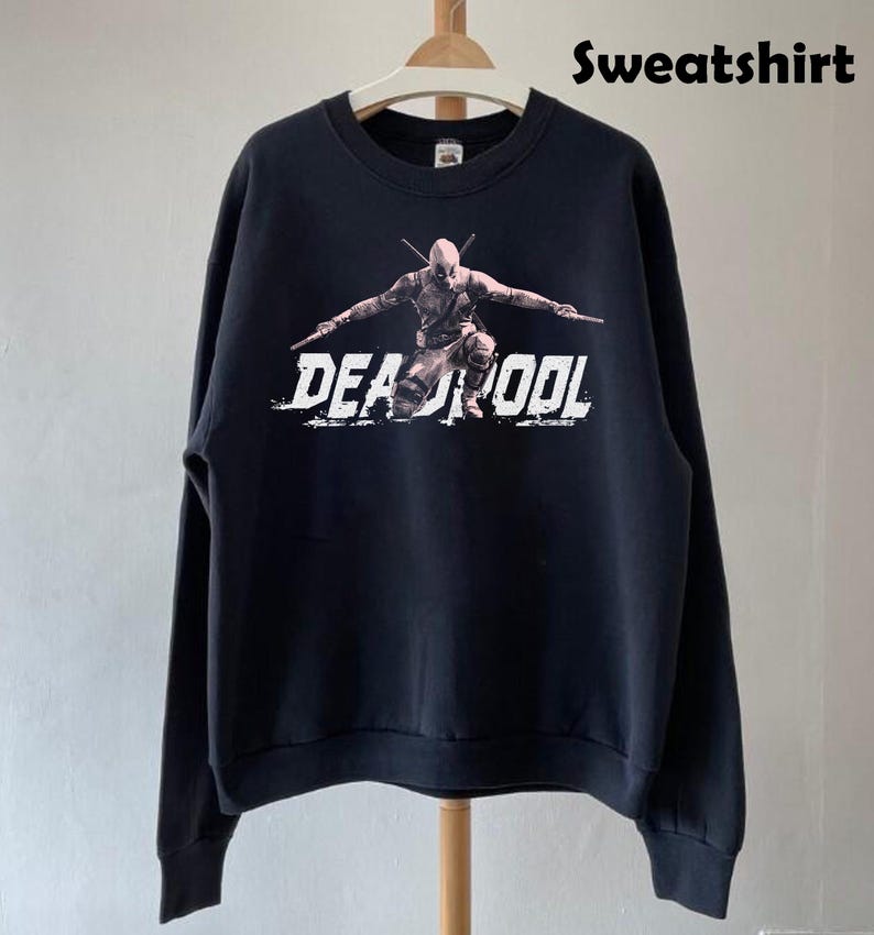 Puede incluir: Sudadera negra con un gr&aacute;fico de Deadpool y la palabra "DEADPOOL" en letras blancas desgastadas. La parte superior de manga larga est&aacute; colgada en una percha de madera.