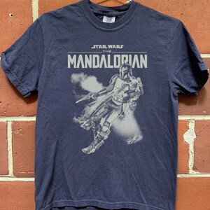 Op de afbeelding: Marineblauw T-shirt met de tekst "STAR WARS THE MANDALORIAN" en een grijze en witte afbeelding van de Mandalorian. Het shirt heeft korte mouwen en een ronde hals. Het T-shirt hangt aan een metalen hanger.