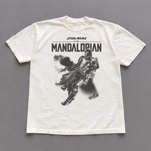 Op de afbeelding: Cr&egrave;mekleurig T-shirt met een zwart-witte afbeelding van The Mandalorian uit Star Wars. De tekst "STAR WARS THE MANDALORIAN" staat boven de afbeelding. Het shirt heeft een ronde hals en korte mouwen.