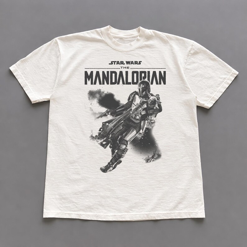 Op de afbeelding: Cr&egrave;mekleurig T-shirt met een zwart-witte afbeelding van The Mandalorian uit Star Wars. De tekst "STAR WARS THE MANDALORIAN" staat boven de afbeelding. Het shirt heeft een ronde hals en korte mouwen.