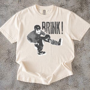 May include: Off-white t-shirt featuring a black and white graphic of a person roller skating, with the word "BRINK!" printed above. The shirt has a crew neck and is made of a soft material.