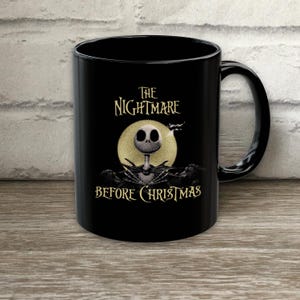 May include: A black ceramic mug featuring a graphic of Jack Skellington from The Nightmare Before Christmas. The design includes a large yellow full moon behind the character, with the text The Nightmare Before Christmas printed in a stylized font.