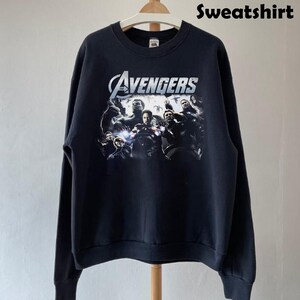 May include: Black sweatshirt featuring a graphic of The Avengers team. The design includes the word "Avengers" in a silver, stylized font above a group of characters. The word "Sweatshirt" is printed in black at the top right.