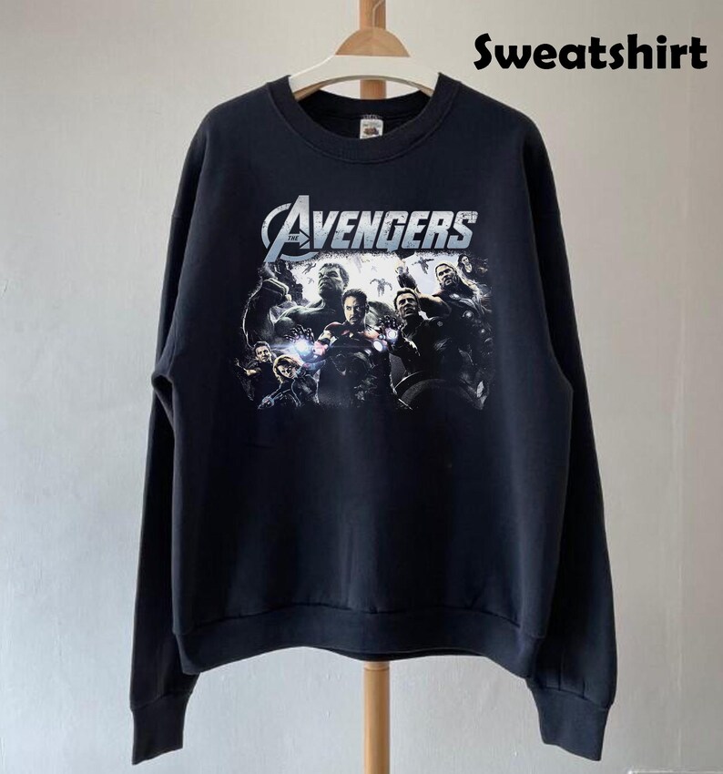May include: Black sweatshirt featuring a graphic of The Avengers team. The design includes the word "Avengers" in a silver, stylized font above a group of characters. The word "Sweatshirt" is printed in black at the top right.