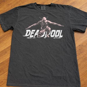 Puede incluir: Camiseta gris oscuro con un gr&aacute;fico de Deadpool arrodillado, sosteniendo dos espadas. La palabra "DEADPOOL" est&aacute; impresa en blanco, con letras desgastadas. La camiseta est&aacute; hecha de un material suave.