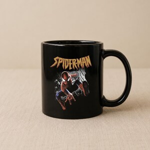 Puede incluir: Una taza de cer&aacute;mica negra con un gr&aacute;fico del personaje Spider-Man en una pose din&aacute;mica. Sobre el personaje, la palabra SPIDER-MAN est&aacute; impresa en una fuente dorada estilizada y desgastada. La taza tiene un asa curva y acabado brillante.