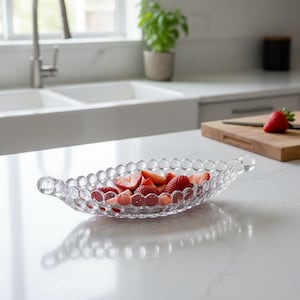 May include: A clear, oval-shaped glass serving dish filled with sliced red strawberries. The dish has a textured, bubble-like design and is placed on a white countertop. The kitchen setting includes a sink, window, and a cutting board.
