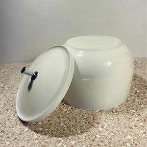 May include: A white, round container with a matching lid. The lid is slightly ajar, revealing the interior. The lid has a chrome handle. The container is on a speckled surface.