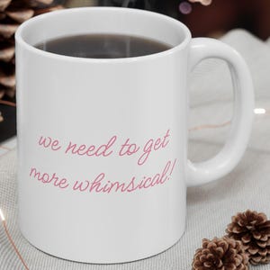 May include: A white ceramic mug with a dark beverage, inscribed with the pink text "we need to get more whimsical!". The mug sits on a light grey surface, with pine cones and a lit candle in the background.