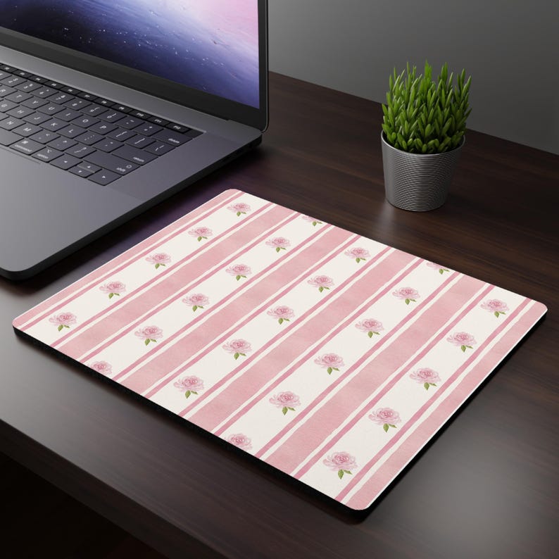 May include: A rectangular mousepad with a pink and white striped pattern. The design features rows of pink roses with green leaves. The mousepad is on a dark brown desk next to a laptop and a small potted plant.