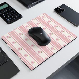 May include: A rectangular mousepad with a pink and white striped pattern and floral accents. A black wireless mouse sits on the pad. Other electronics, including a calculator and phone, are in the background.