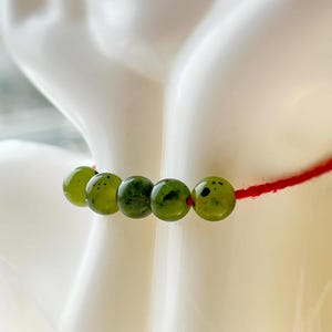 Könnte beinhalten: Ein Armband mit fünf grünen, gesprenkelten Perlen auf einer dünnen roten Schnur. Die Perlen sind eine Mischung aus hell- und dunkelgrün, mit glatter Oberfläche. Das Armband wird vor weißem Hintergrund präsentiert.