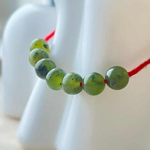 Könnte beinhalten: Nahaufnahme eines Armbands mit sieben grünen Jadeperlen auf einer roten Schnur. Die Perlen sind rund und gesprenkelt. Der Hintergrund ist unscharf, mit einem weißen Objekt im Hintergrund.