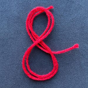 Puede incluir: Hilo rojo brillante en forma de ocho sobre un fondo de tela azul oscuro. La textura del hilo es visible y los extremos son claros. Ideal para proyectos de manualidades o temas textiles.