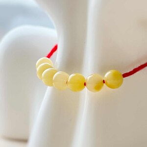 May include: A bracelet with pale yellow, spherical beads threaded on a red cord. The bracelet is shown against a white backdrop, emphasising the colours and design. The beads appear to be made of a translucent material.