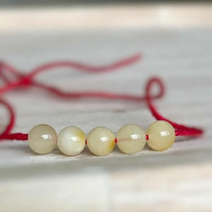 May include: A bracelet with five spherical, pale yellow beads threaded on a red cord. The beads have a glossy finish and are evenly spaced. The red cord is visible on either side of the beads.