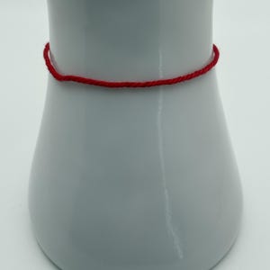 May include: A thin, bright red bracelet encircles a white, vase-shaped object. The bracelet appears to be made of twisted thread or cord. The vase has a smooth, glossy finish and a simple, elegant design.