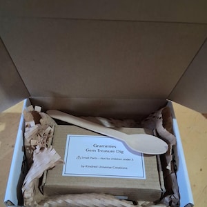 May include: A cardboard box containing a small brown box, packing paper, and a wooden spoon. A label on the small box reads "Grammies Gem Treasure Dig" with a warning about small parts. The box is open, revealing the contents.