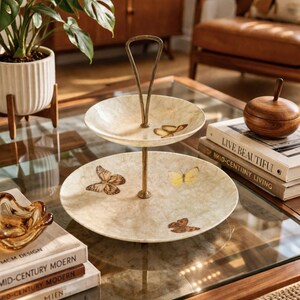 May include: Two-tiered serving tray with a gold-tone handle and stand. The off-white plates are decorated with butterfly illustrations in shades of brown and yellow. The tray is displayed on a glass-top table.