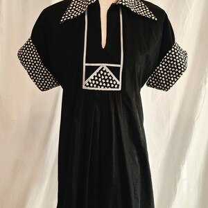 May include: A black dress with short sleeves and a pointed collar. The collar, sleeve cuffs, and a rectangular chest panel feature a white floral pattern on a black background. The dress has a split neckline and vertical pleats below the chest panel.