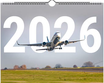 Può includere: Calendario 2026 con una fotografia di un aereo passeggeri che decolla da una pista. L'aereo è bianco e blu, con l'anno 2026 in grandi numeri bianchi sullo sfondo. Il calendario ha una rilegatura a spirale.