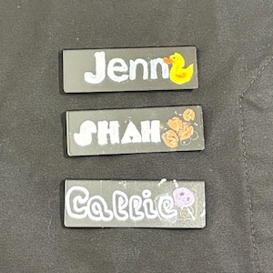 May include: Three rectangular name tags with white lettering on a gray background. The top tag says "Jenn" with a yellow duck graphic. The middle tag says "Shah" with a brown graphic. The bottom tag says "Carrie" with a purple graphic.