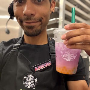 May include: A clear plastic cup filled with a layered purple and orange beverage, topped with a green straw. The cup is held by a person wearing a black apron with the Starbucks logo and the text "Coffee Master". A name tag reads "ADRIAN".