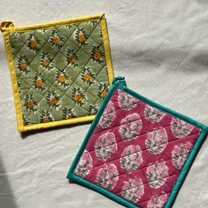 May include: Two square quilted pot holders. One is green with yellow trim and a floral pattern. The other is pink with teal trim and a floral pattern. Both have a loop for hanging.