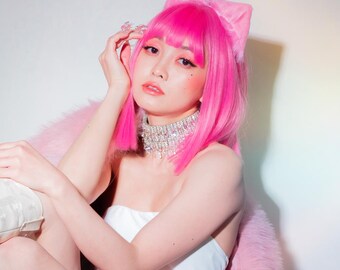 May include: A woman with pink hair and cat ears sits on a white chair with a white table in front of her. She is wearing a white top, a pink skirt, and white boots. The chair and table are both ornate and have a white, lacy design.