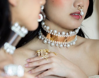 May include: Two women wearing pearl and rhinestone chokers. One woman is wearing a gold ring with rhinestones. Both women are wearing white tops with lace trim.